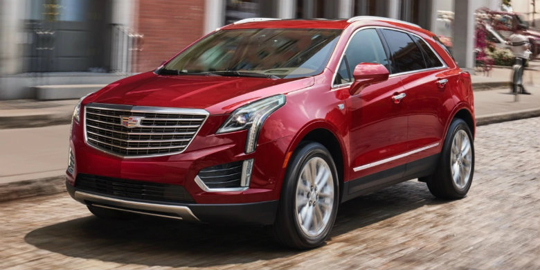 Front exterior view of a red, used 2019 Cadillac XT5 driving on a brick city street with a buildings in the background.