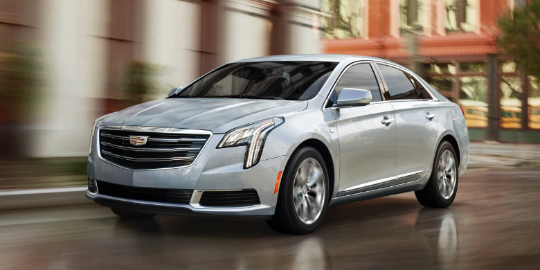 Front exterior view of a silver used 2019 Cadillac XTS drivingo n a city street with buildings and businesses in the background.