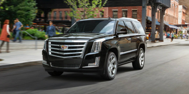 Front exterior view of a black, used 2019 Cadillac Escalade driving on a city street with people walking and businesses in the background
