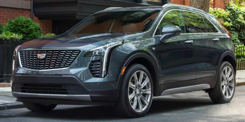 Front exterior view of a dark teal, used 2019 Cadillac XT4 parked on a city street with a brick building and hedges in the background.