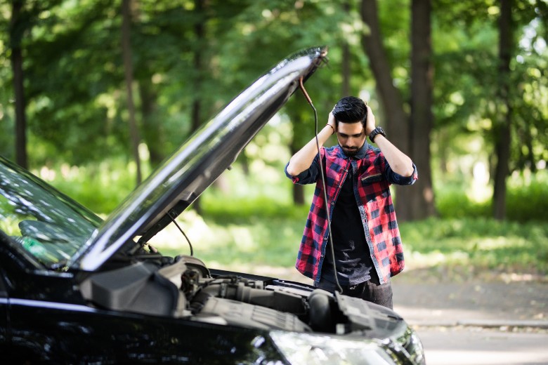 Man with hands on head beside broken down car with hood open